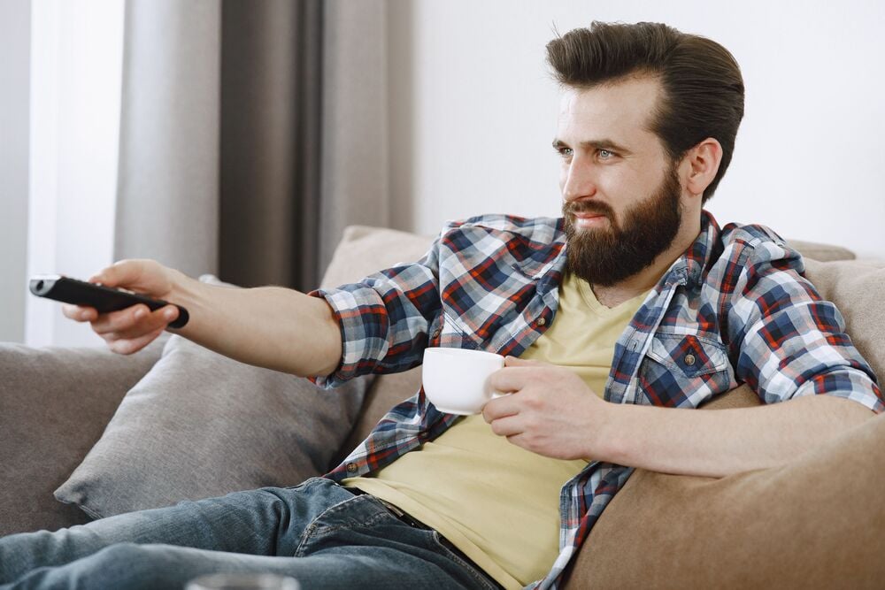 A bearded man in a plaid shirt sits on a couch, holding a white mug in one hand and pointing a remote control with the other, looking at something off-camera.