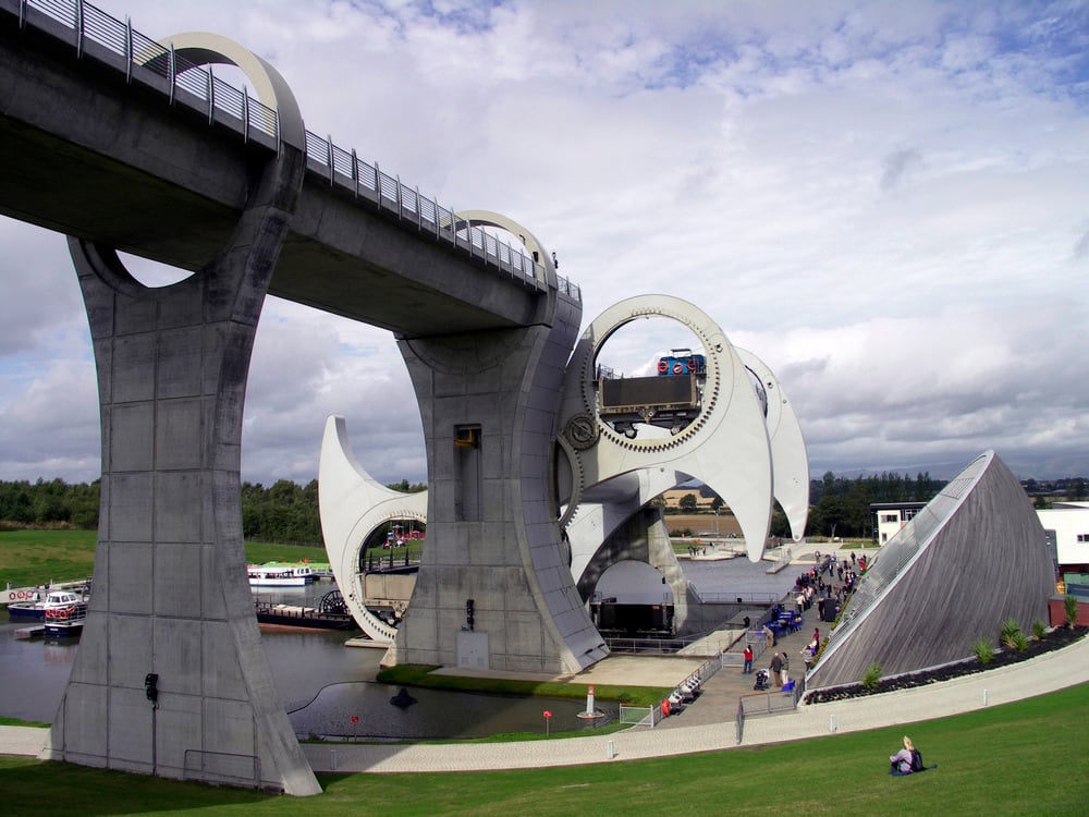 A large rotating boat lift with curved concrete arms and walkways connects two water levels, surrounded by green grass, people, and boats, under a cloudy sky.