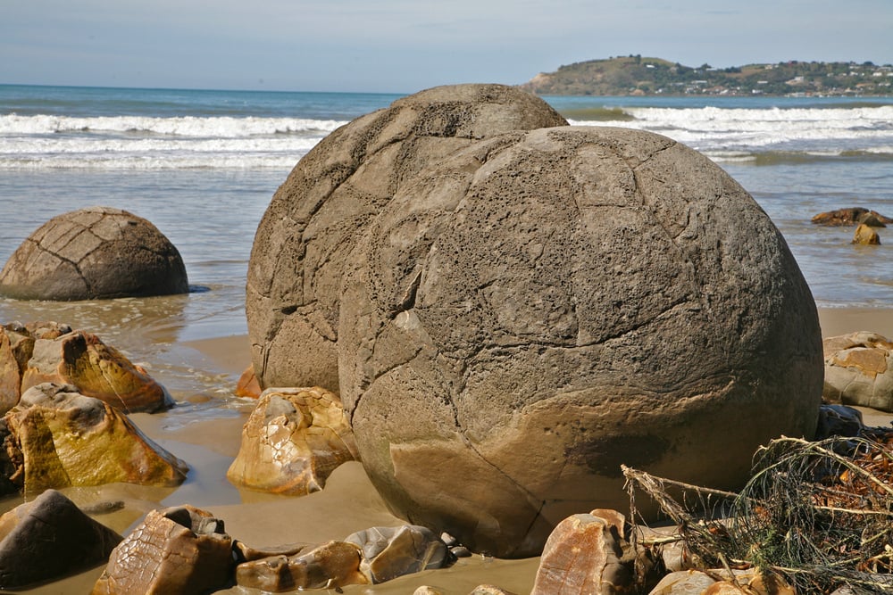 Large, round boulders sit on a sandy beach with waves in the background and rocky hills in the distance under a partly cloudy sky.