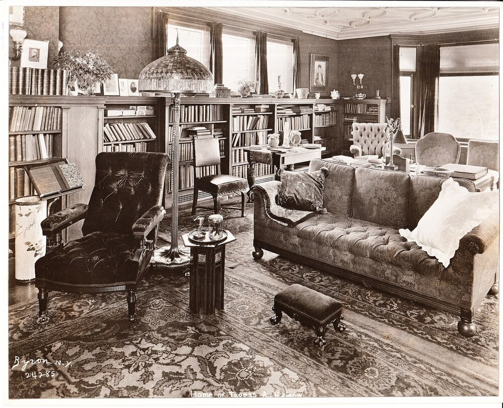 A vintage sitting room with bookshelves, armchairs, a sofa, and large windows.