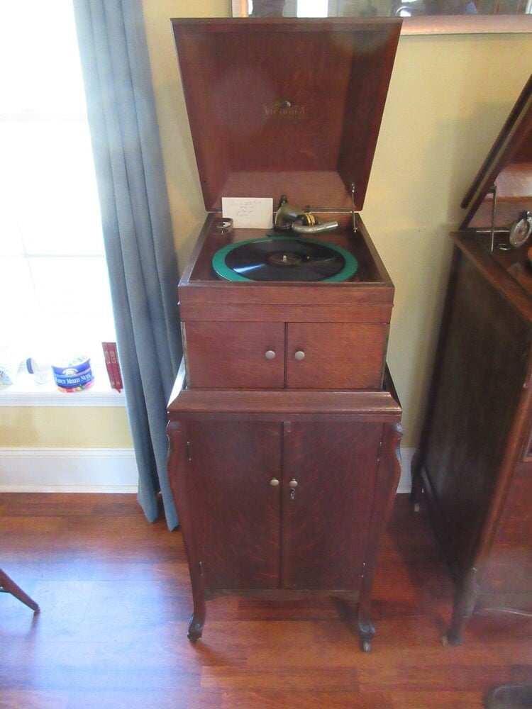 An antique wooden Victrola record player with its lid open, showing the turntable and tonearm, stands on hardwood floor next to a window with light blue curtains.