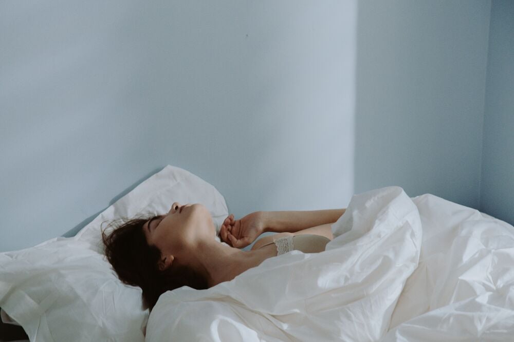 A person lies in bed on white sheets with eyes closed, resting against a light blue wall.