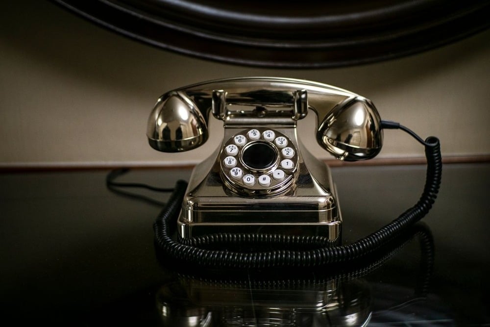 A shiny, vintage rotary dial telephone with a coiled cord sits on a dark reflective surface, with a neutral-colored wall and a framed mirror in the background.