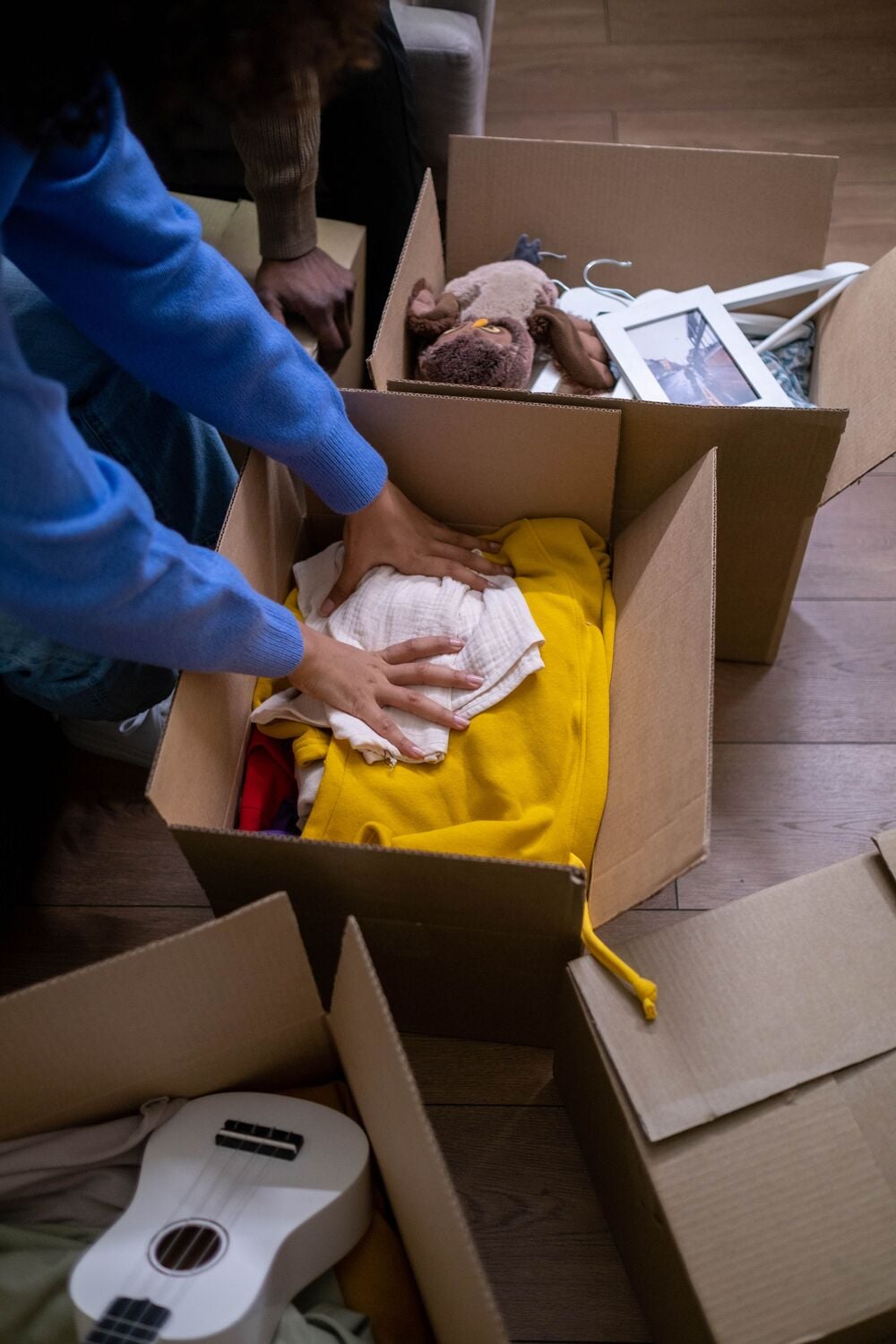 A person packs clothes into a cardboard box surrounded by other boxes containing a ukulele, stuffed animal, and a photo frame on a wooden floor.