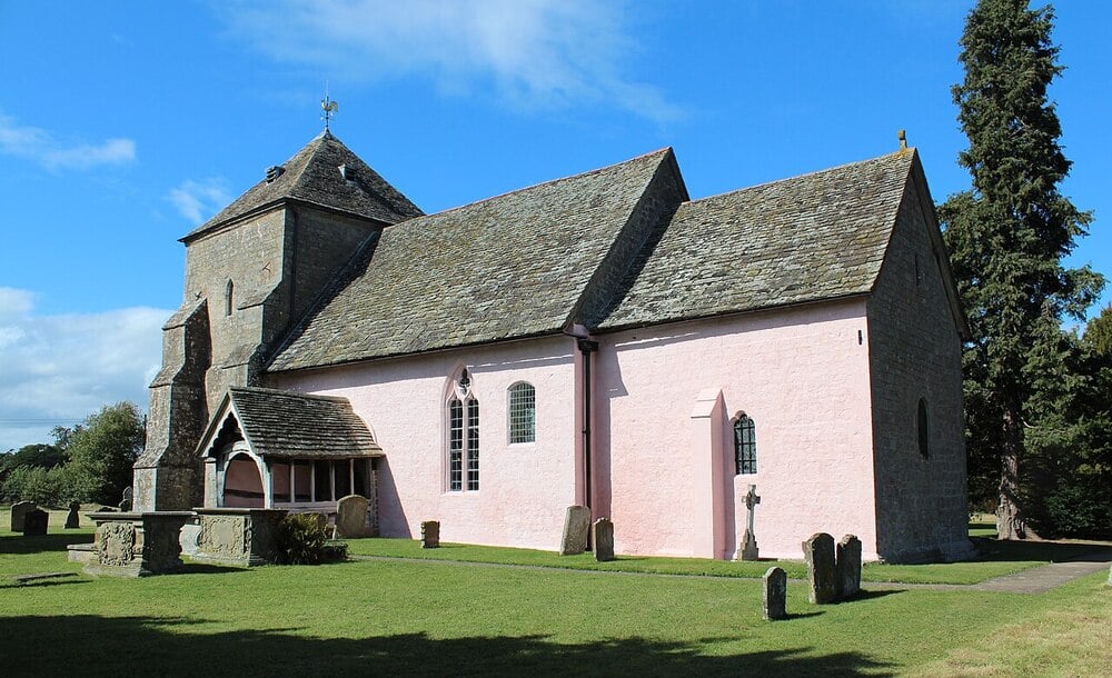 A historic stone church with a pink exterior, arched windows, and a shingled roof stands in a grassy churchyard with gravestones under a blue sky.