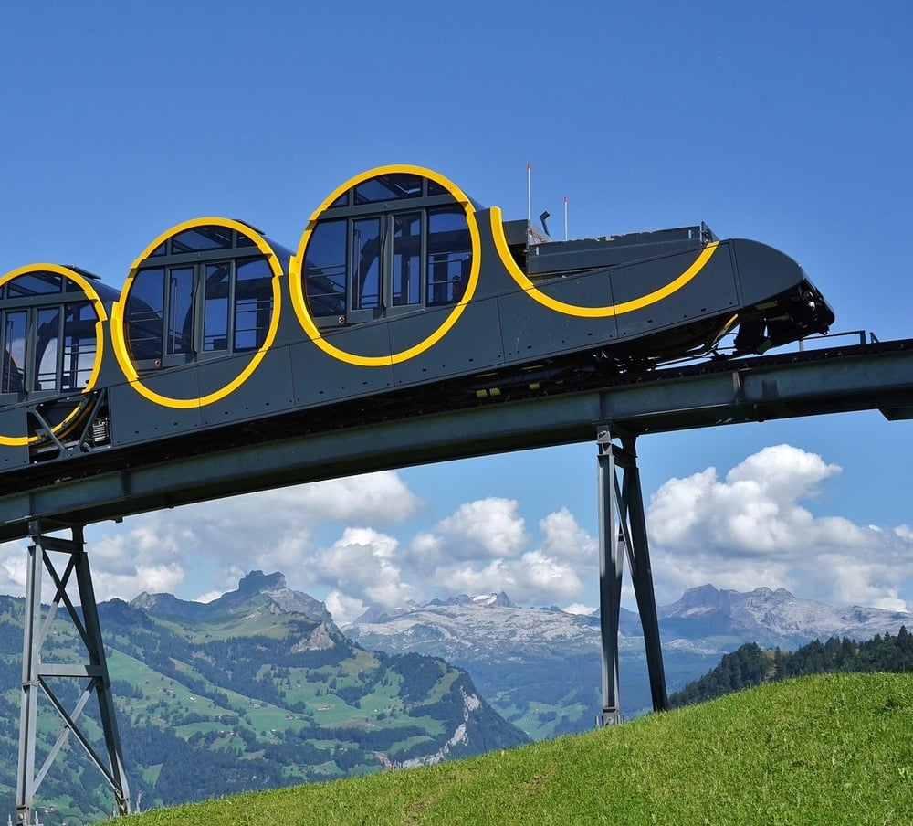 A modern, circular-funicular railway car with yellow accents travels uphill on a track through green hills, with mountains and a partly cloudy blue sky in the background.