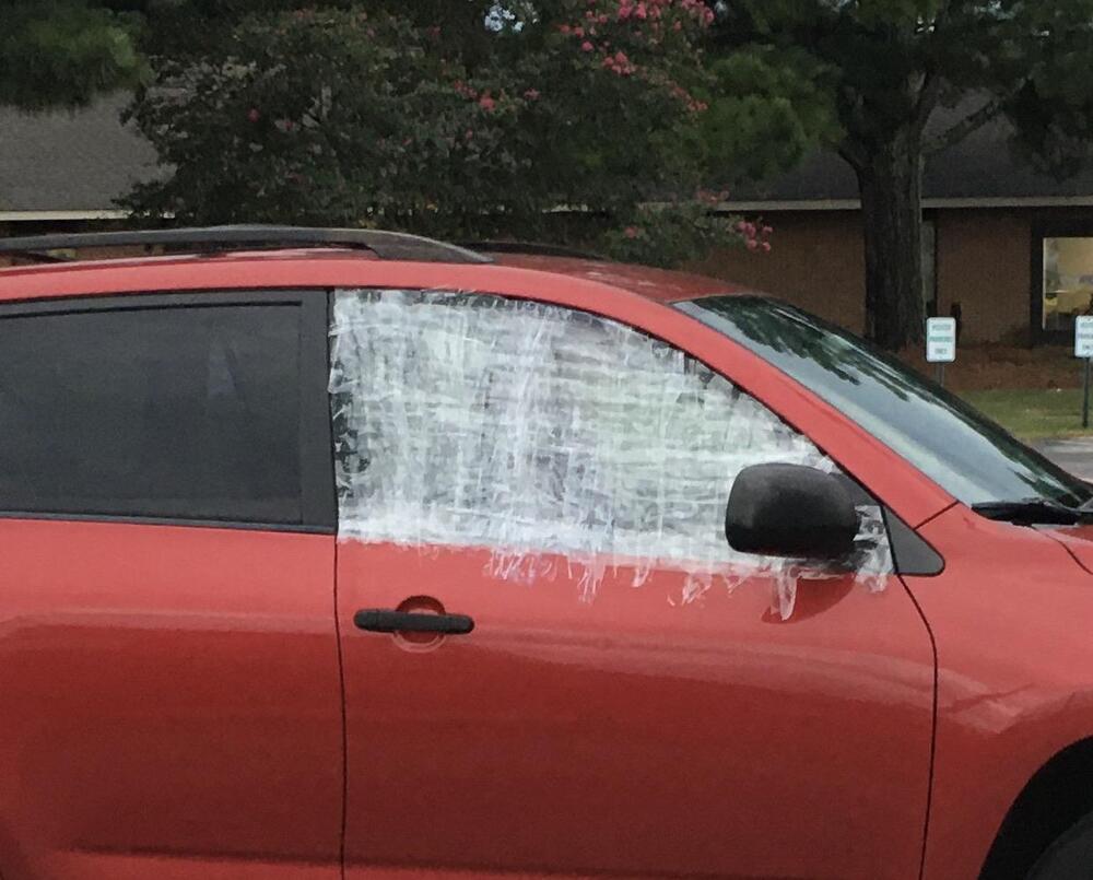 A red car with plastic wrap taped over the driver-side window, possibly as a temporary replacement for broken glass. Trees and a building are visible in the background.