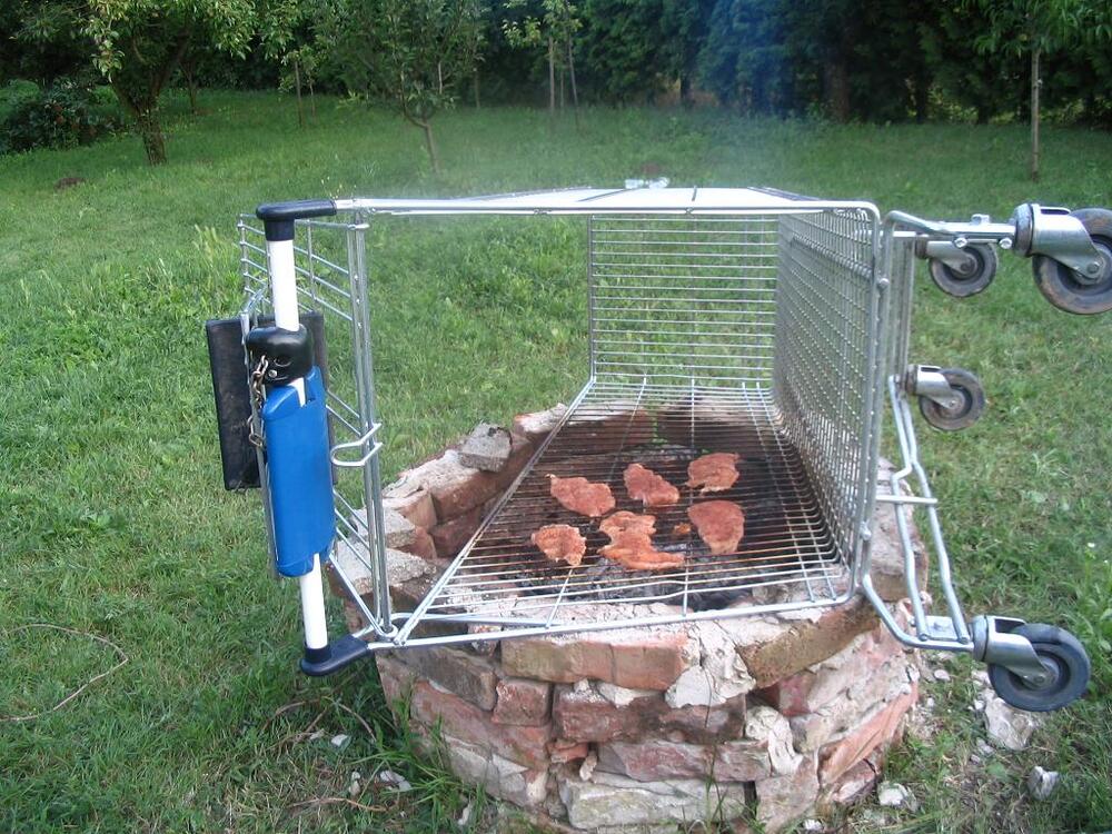 A shopping cart turned on its side is being used as a grill over a brick fire pit in a grassy yard, with several pieces of meat cooking inside.