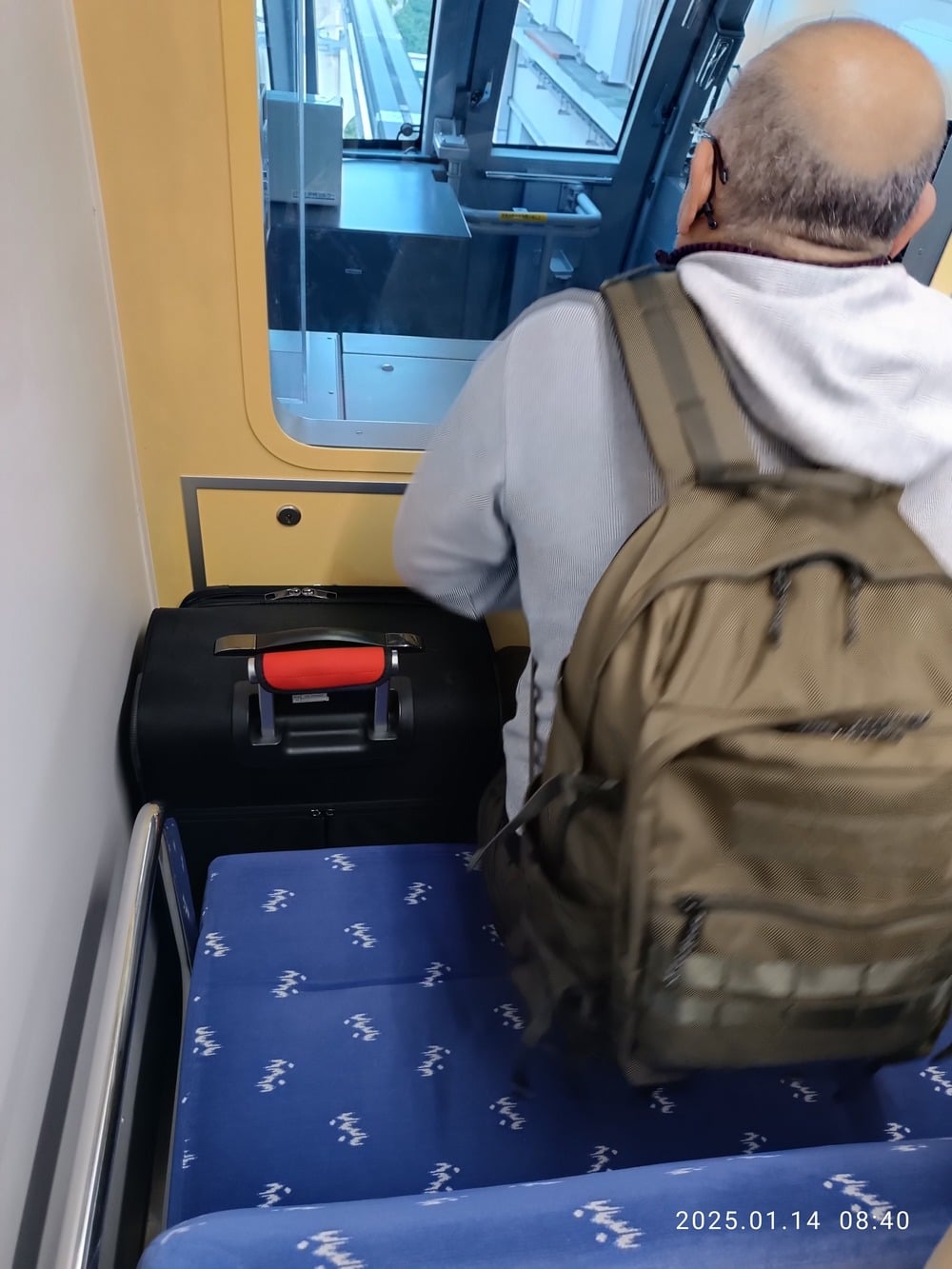 A person with a backpack stands beside a suitcase on a blue patterned seat, facing a window on a train or tram. The timestamp on the photo reads “2025.01.14 08:40.”