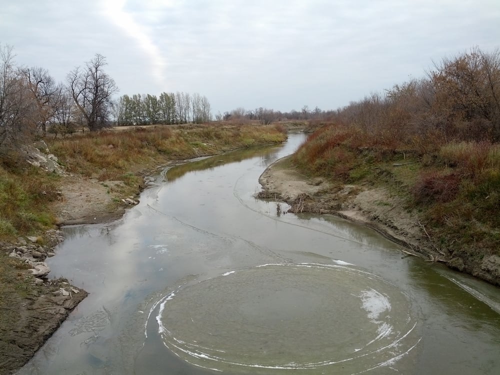 A calm river with partially frozen water forms circular ice patterns. Bare trees and dry grass line the banks under a cloudy sky. The landscape appears cold and quiet in late autumn or early winter.