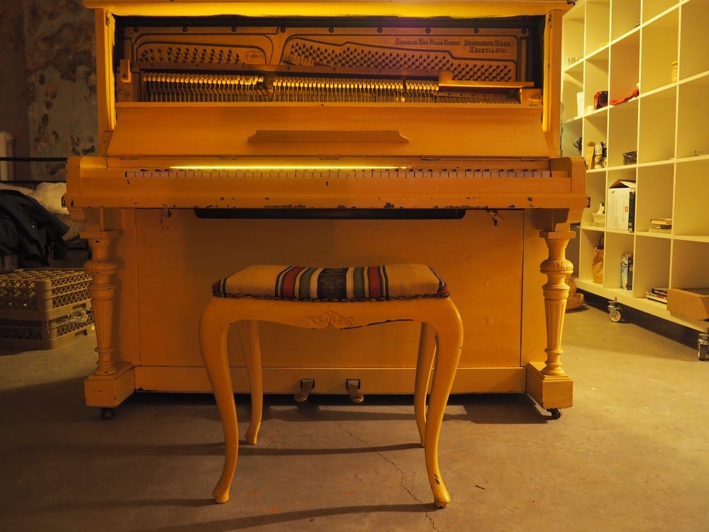 A yellow upright piano with an open top exposing its strings stands behind a matching yellow bench with a striped cushion in a warmly lit room.