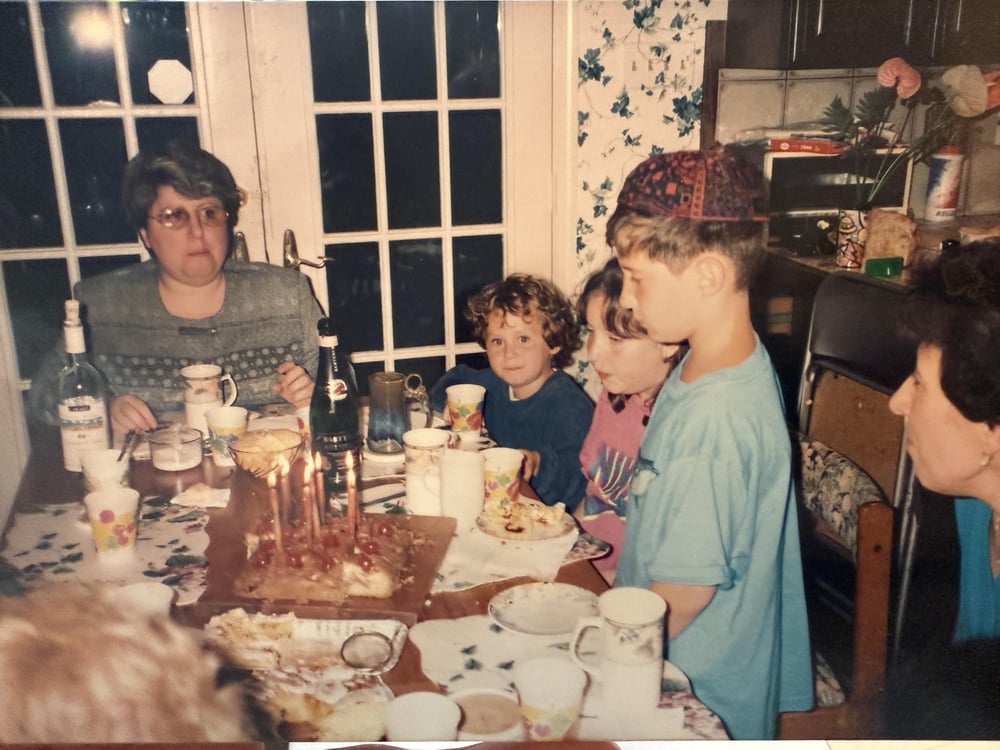 Children and adults sit around a birthday table with cake, food, and drinks.