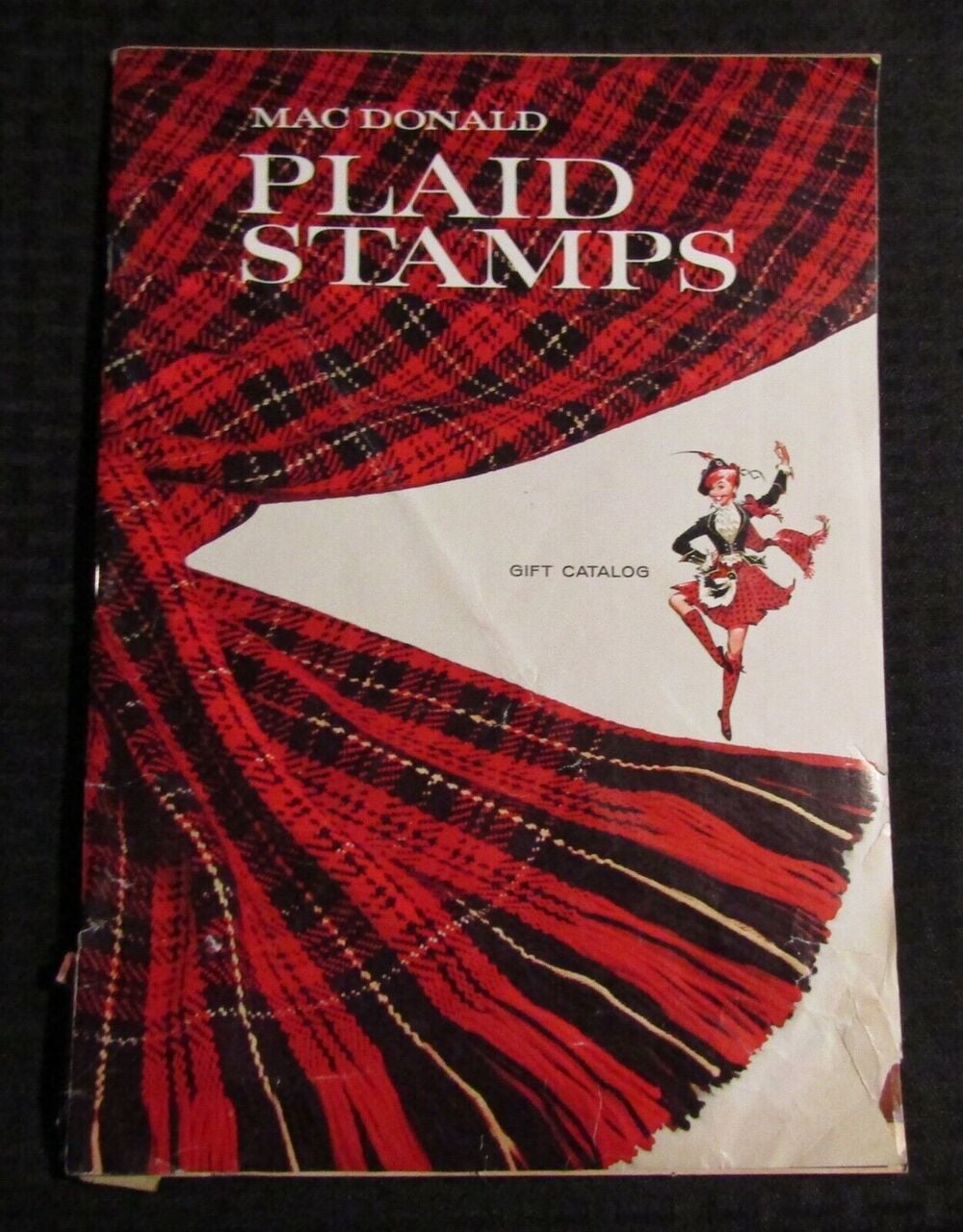 Cover of a MacDonald Plaid Stamps gift catalog, showing a large red plaid curtain pulled back to reveal an illustration of a woman in a Scottish outfit dancing.