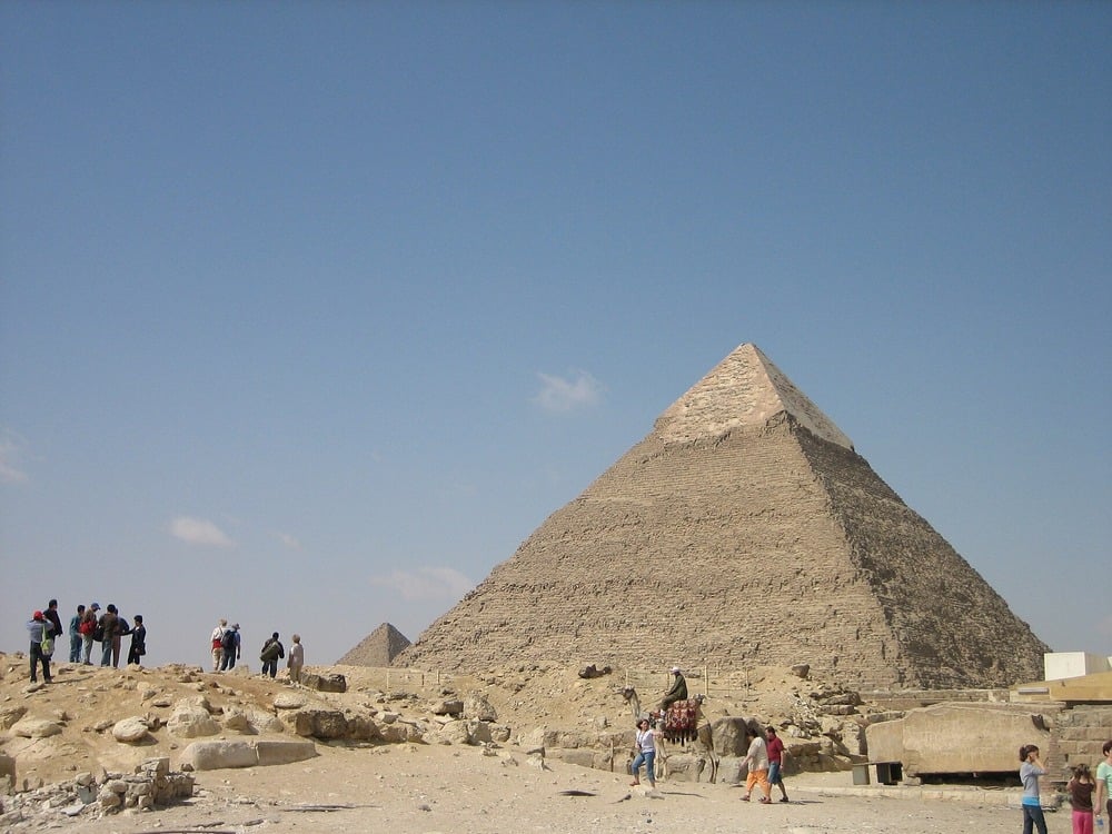 The Great Pyramid of Giza under a clear sky, with groups of tourists walking and taking photos near its base.