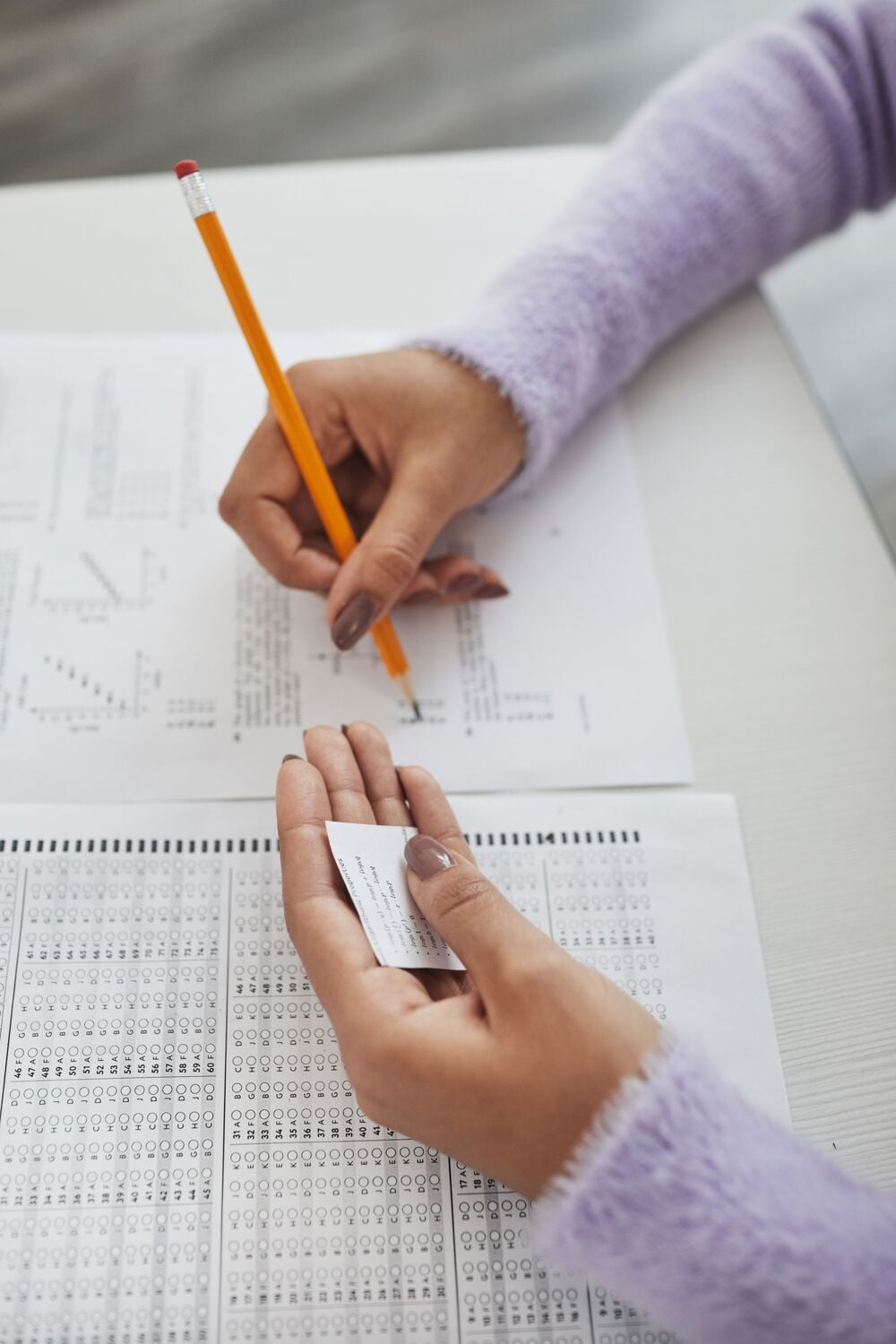 A person wearing a lavender sweater fills out a multiple-choice exam with a pencil while secretly holding a small cheat sheet in their left hand.
