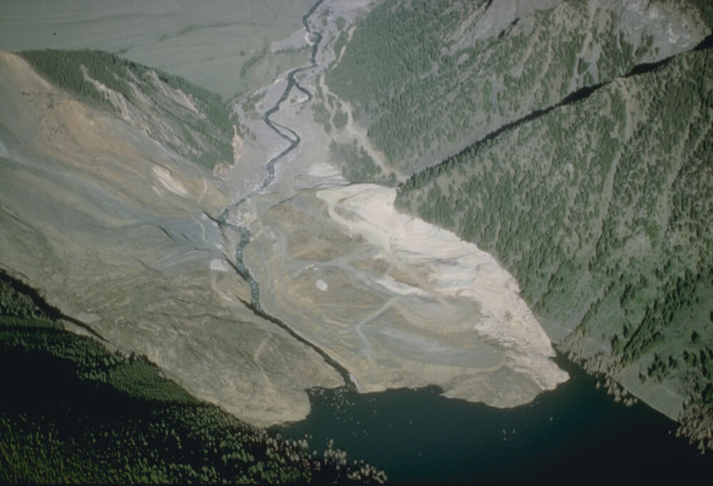 Aerial view of a river flowing through a valley into a lake, with visible sediment deposits and surrounding forested and mountainous terrain.