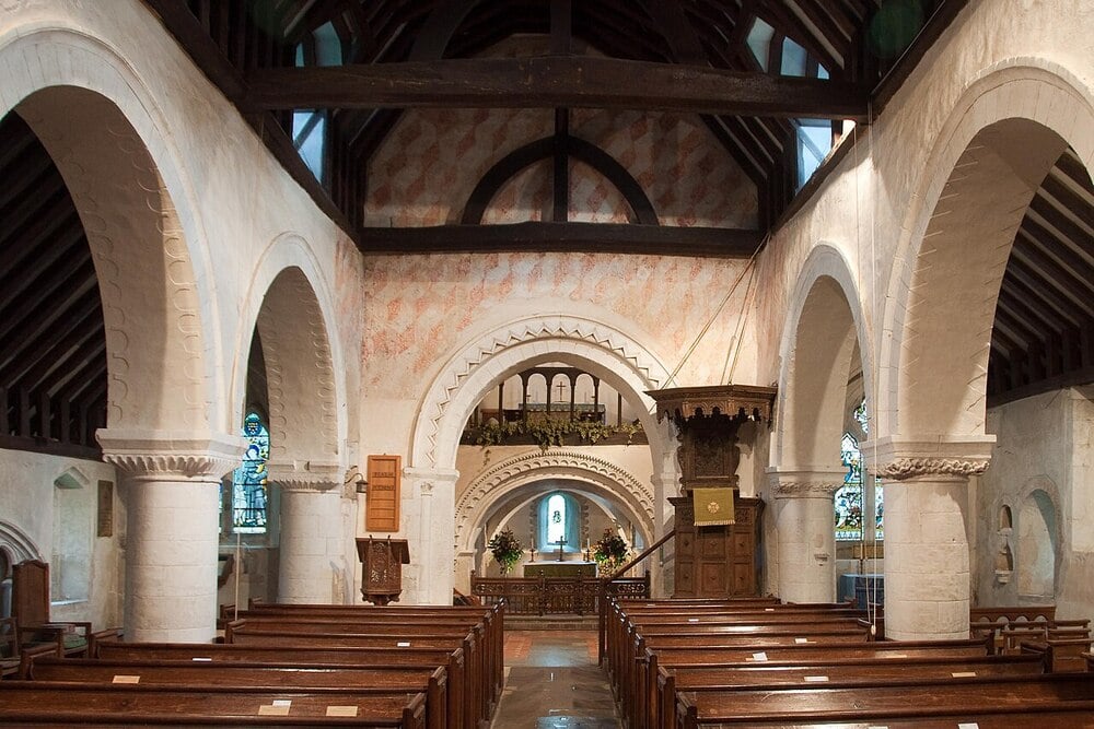 Interior of a historic stone church with wooden pews, arched columns, a painted chancel arch, stained glass windows, and exposed wooden ceiling beams.