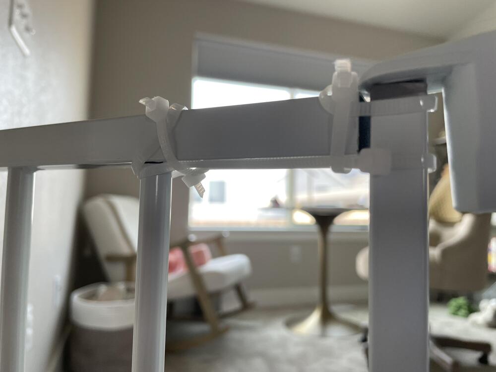 Close-up of a baby gate secured with white zip ties, with a blurred background showing a rocking chair, table, and window in a softly lit room.