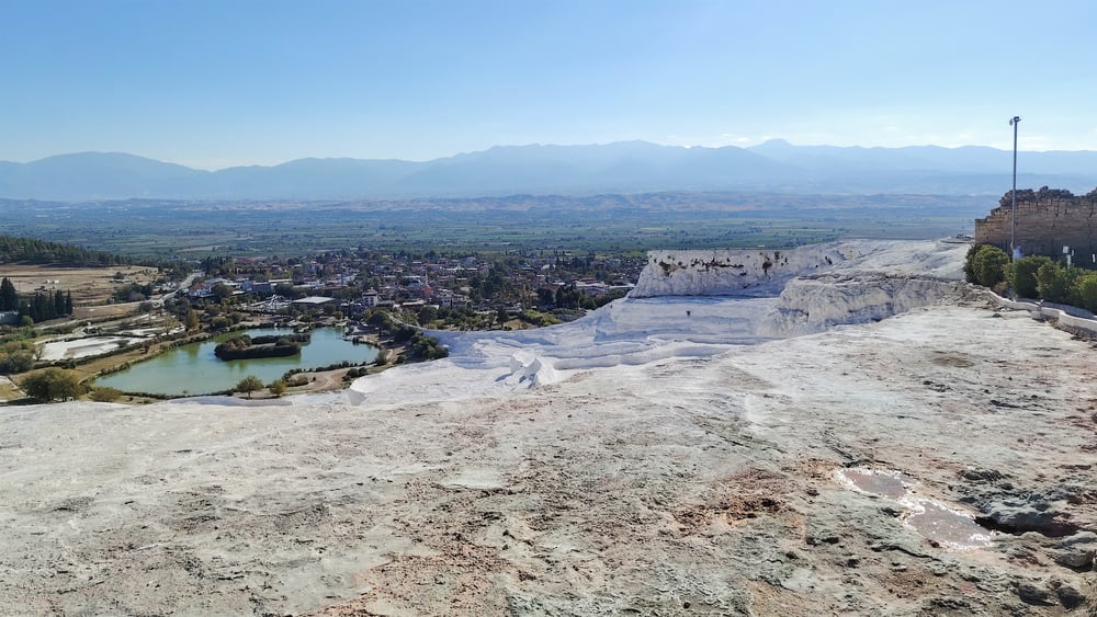 White travertine terraces at Pamukkale, Turkey, overlooking a small lake and village with distant green fields and mountains under a clear blue sky.