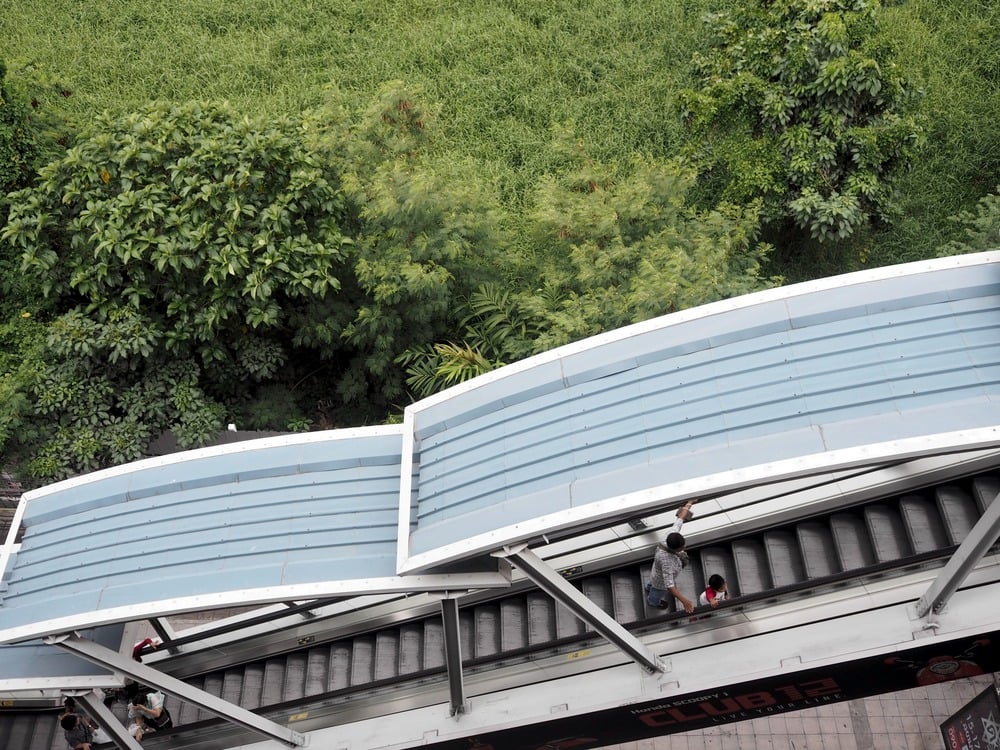 Aerial view of covered outdoor escalators with people riding, bordered by lush green trees and grass.