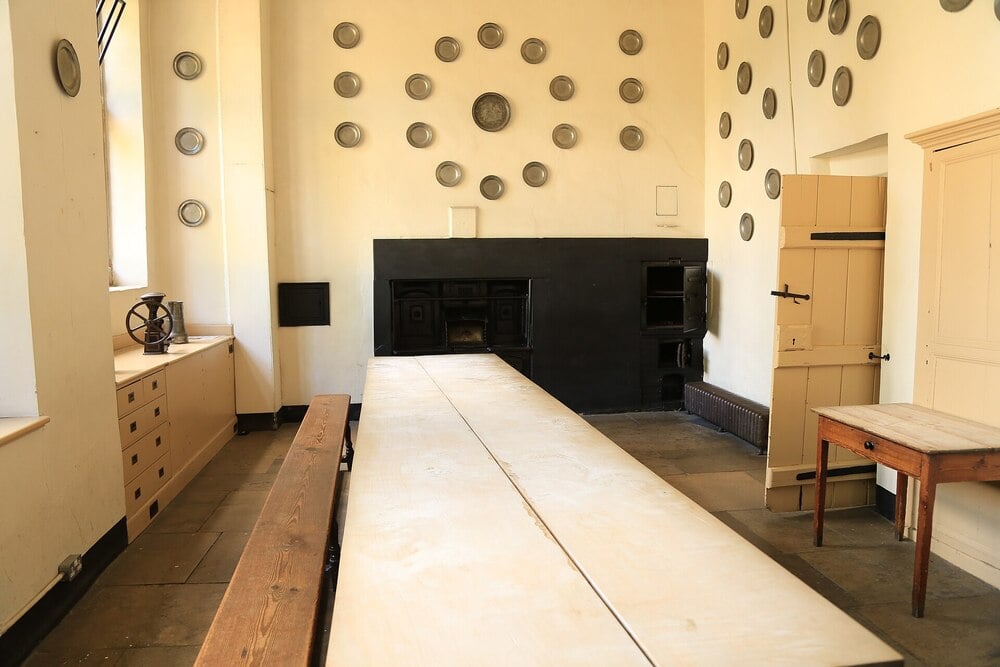 A historic servant's hall with a long wooden table, benches, and a fireplace.