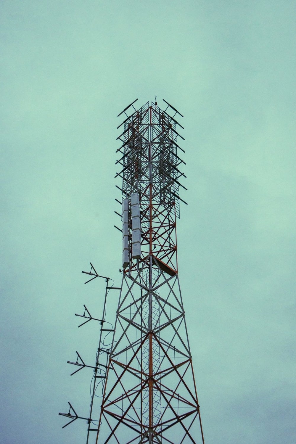 A tall metal communication tower with multiple antennas and panels extends into a cloudy, pale blue sky.