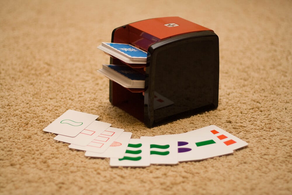 A card shuffler sits on a carpet next to a fan of playing cards displaying various shapes and colors, including squiggles, ovals, and rectangles in green, purple, and red.