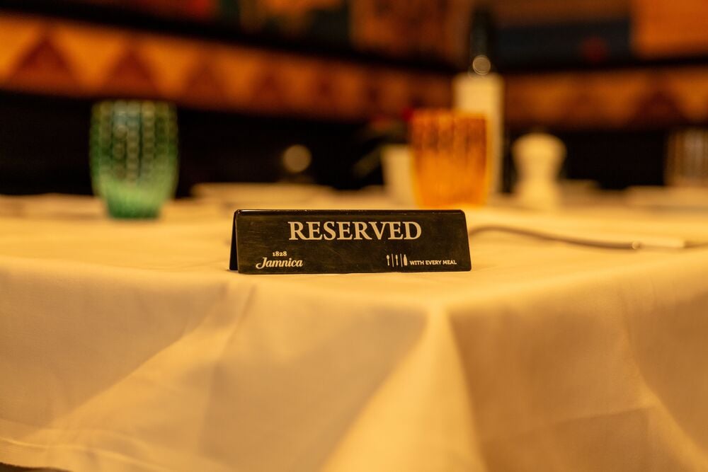 A “Reserved” sign sits on a white tablecloth in a restaurant, with blurred colorful glasses and a bottle in the background.