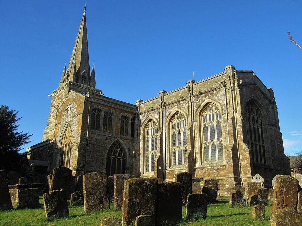 A historic stone church with a tall spire stands under a clear blue sky, surrounded by old, weathered gravestones in a grassy churchyard.
