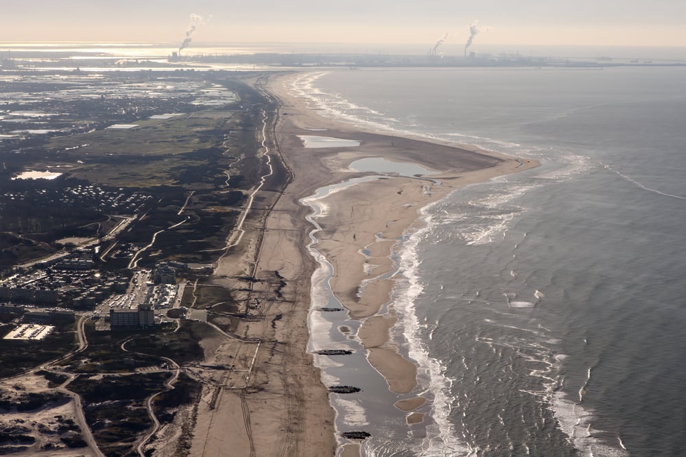 Aerial view of a sandy coastline with gentle waves, buildings near the shore, green areas inland, and factory smokestacks in the distant background emitting smoke.