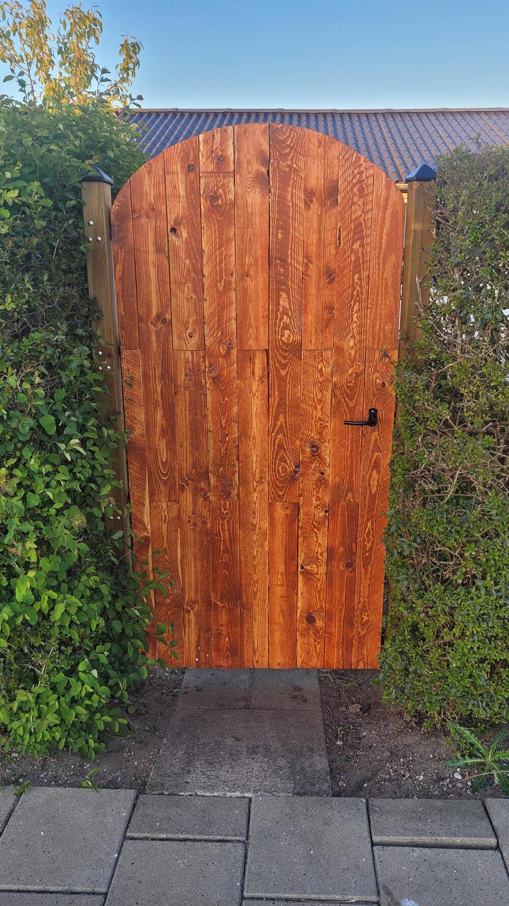 A wooden garden gate with a curved top stands between green hedges, with a stone path leading up to it. The gate has a natural wood finish and a small black latch on the right side.