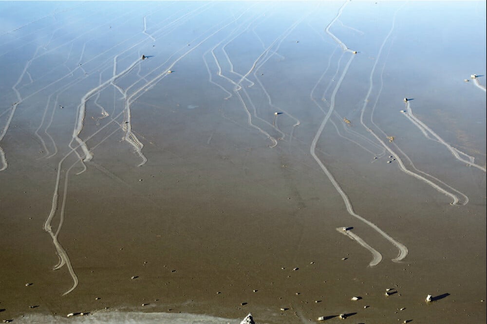 Large rocks on a flat, brown surface with long trails behind them, showing paths where rocks have slowly moved across the ground, under a pale sky reflection.