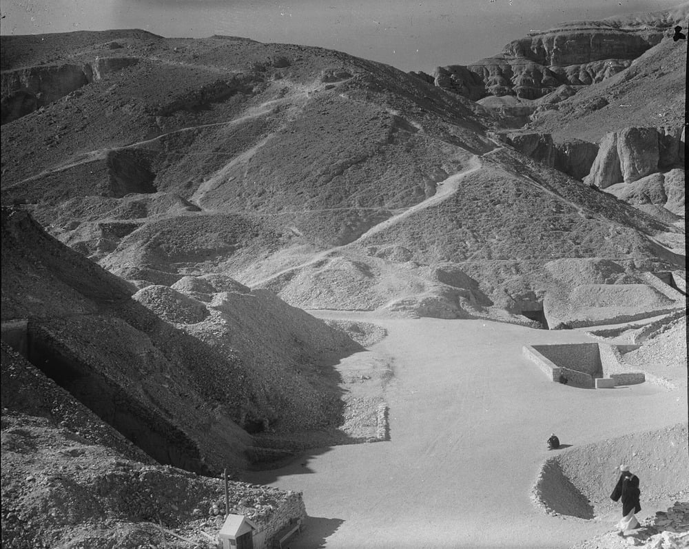 Black and white photo of a rocky, barren valley with winding paths on hillsides. Two people stand on a wide dirt area; one appears to be photographing the landscape.