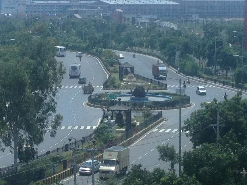 Aerial view of a busy roundabout with a large fountain in the center, surrounded by trees, vehicles, and buildings in the background.
