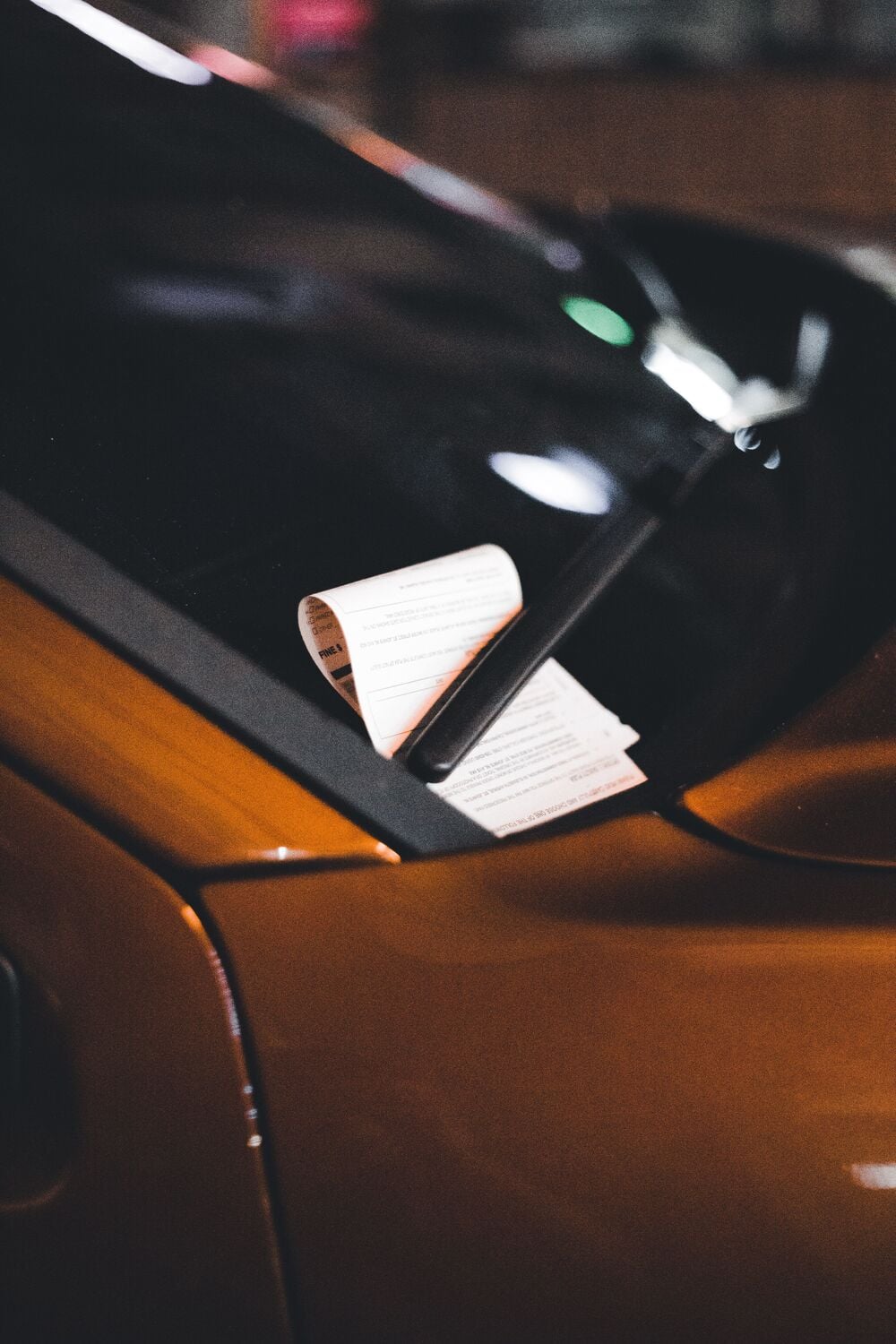 A close-up of a parking ticket tucked under the windshield wiper of an orange car, photographed at night.
