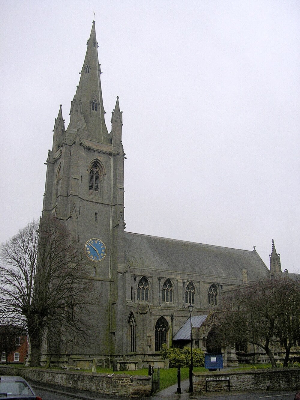A large, gray stone church with a tall clock tower and spire stands near leafless trees on a cloudy day. A blue sign and a car are visible in front.