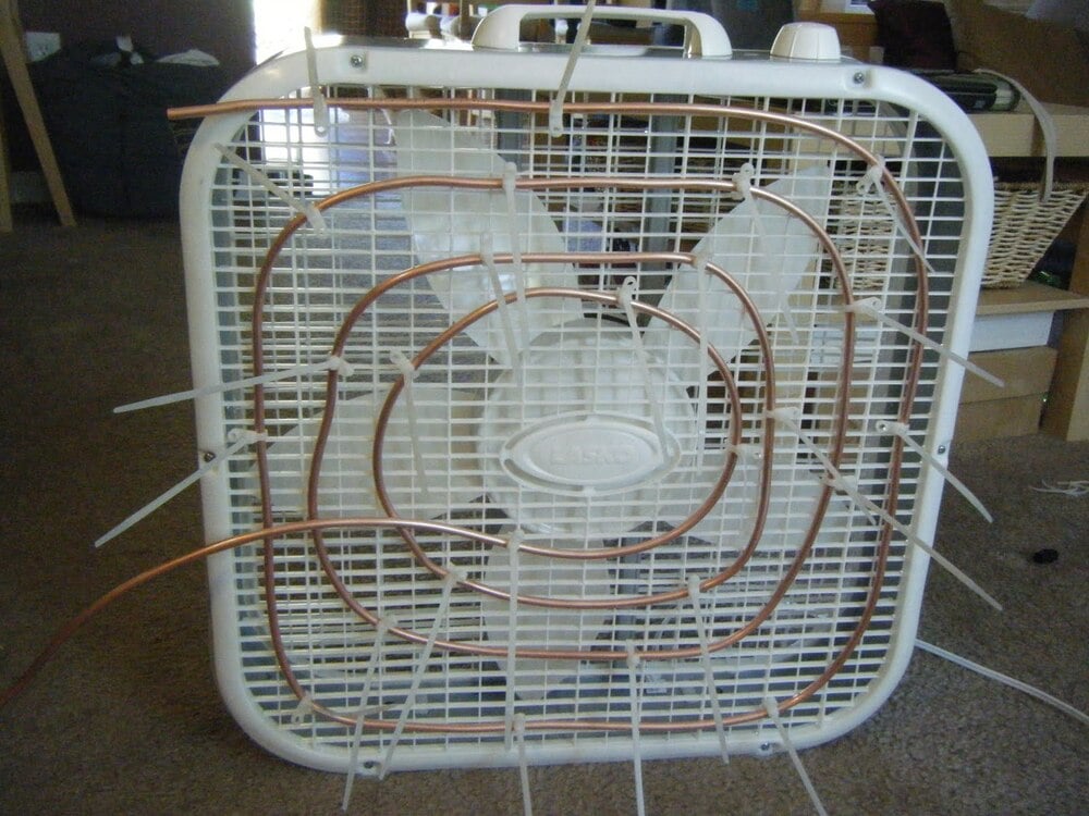A box fan with copper tubing and zip ties attached to the front grill, placed on a carpeted floor indoors.
