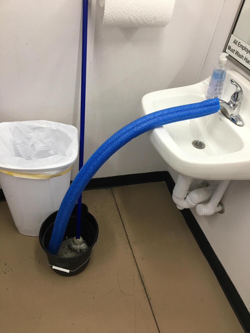 A blue pool noodle bridges a small sink to a mop bucket, directing water into the bucket. A mop and trash can are nearby in a restroom.