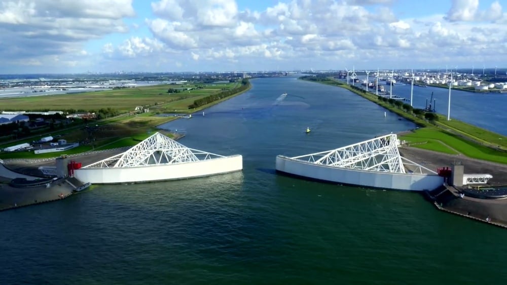 Aerial view of Maeslantkering storm surge barrier in the Netherlands, with massive white gates partially closed over a wide waterway, surrounded by green fields and industrial structures.