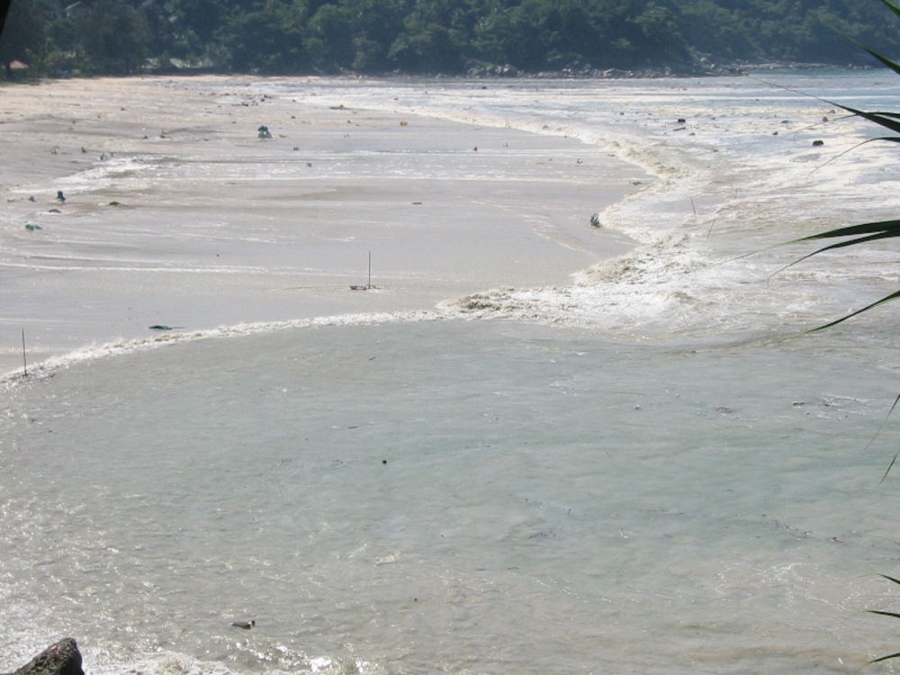 A strong ocean current flows across a sandy beach, creating a visible rip in the waves, with trees and greenery visible in the background.