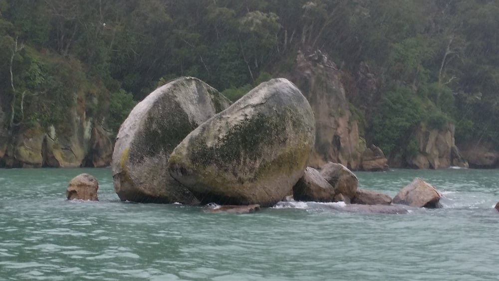 Large, rounded boulders covered in moss sit in turquoise water near the shore, with a forested and rocky background under an overcast sky.