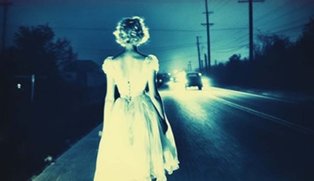 A woman in a white dress stands on a deserted road at night, illuminated by an approaching car’s headlights, with power lines and trees in the background.