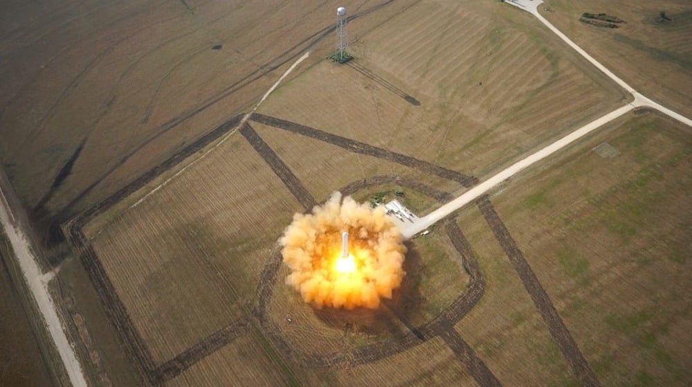 A rocket launches vertically from a circular pad, creating a bright orange fireball and smoke plume, surrounded by open fields and dirt roads, as seen from above.