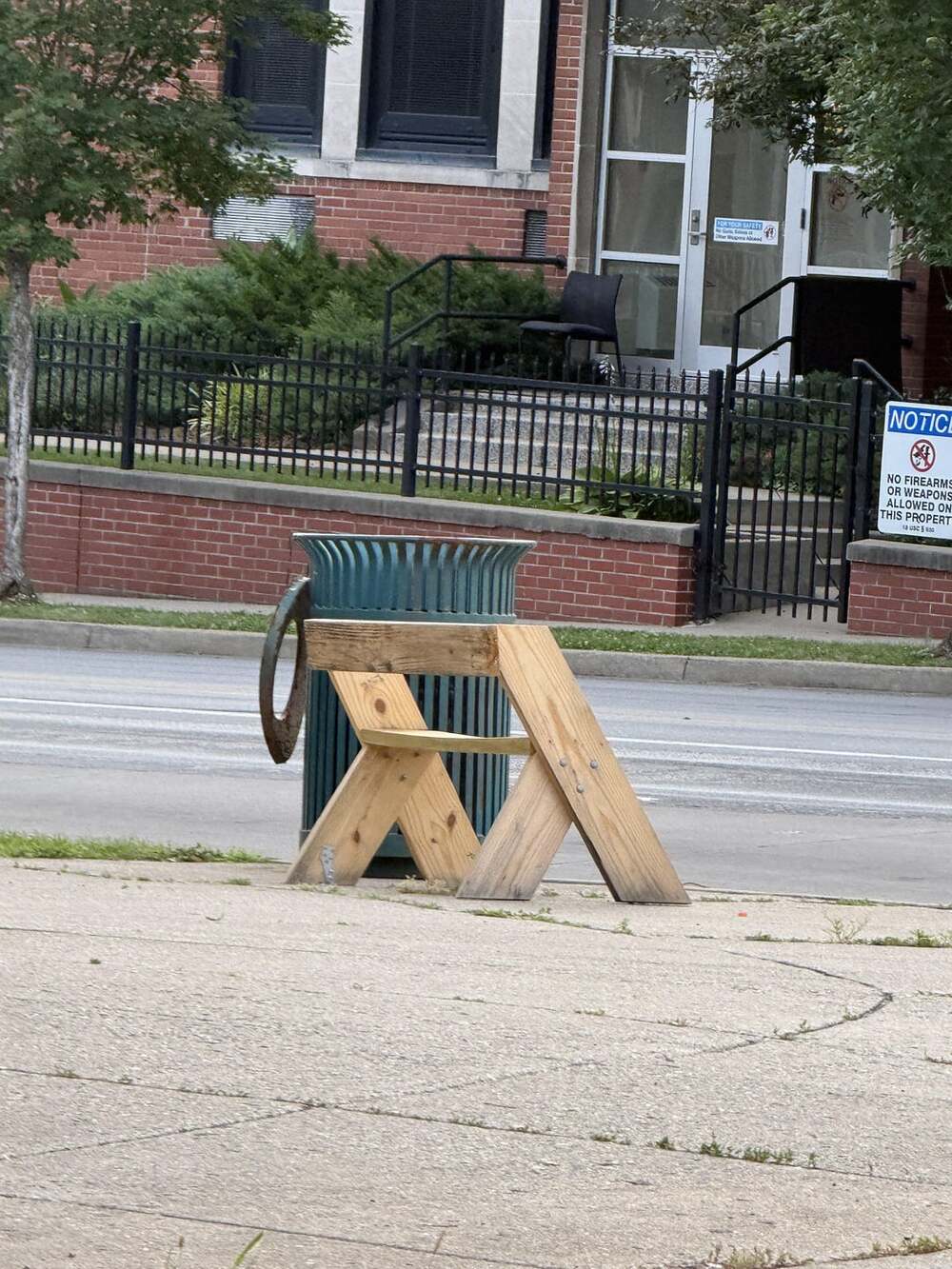 A small wooden bench is positioned on a sidewalk near a green trash can, with a street, building, and some greenery in the background.