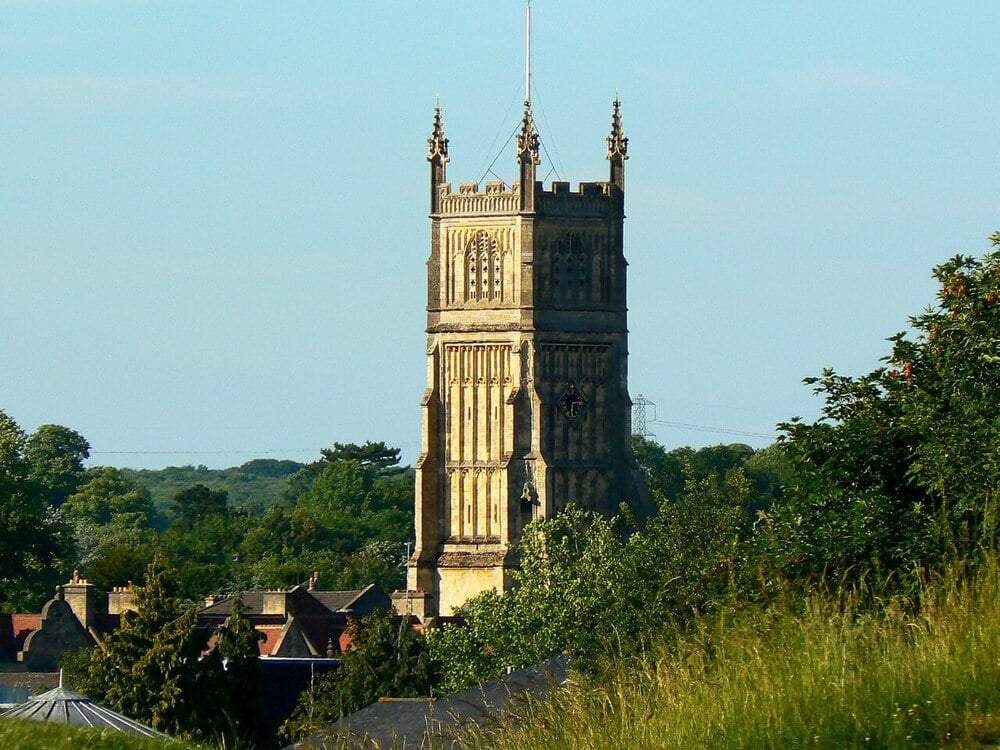A tall, ornate stone church tower rises above rooftops and green trees under a clear blue sky on a sunny day.