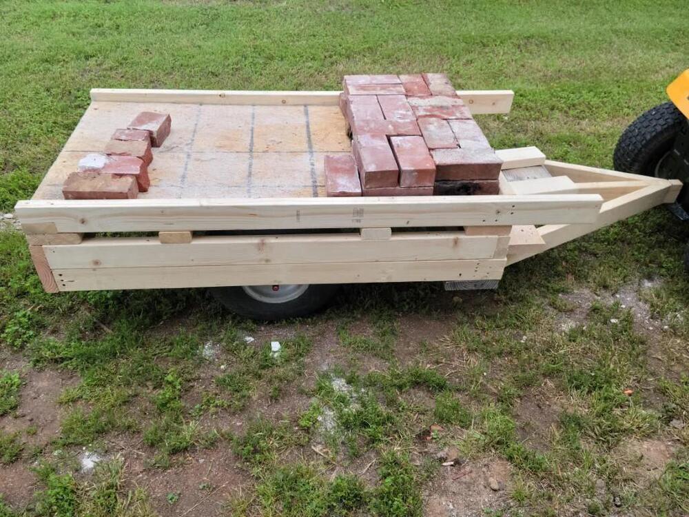 A wooden flatbed cart with a wheel in the center is loaded with red bricks and sits on grass. The cart has wooden slats and is attached to a vehicle on the right side of the image.