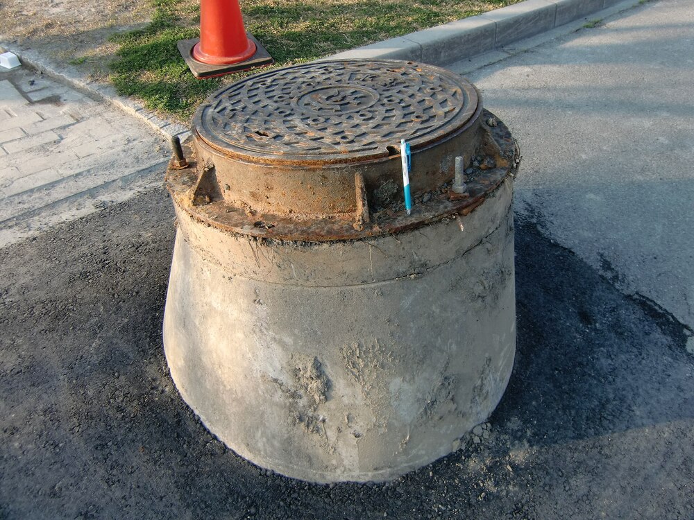 A raised manhole with a rusted cover sits above street level on a patch of asphalt near a curb, with a traffic cone and grass visible in the background.