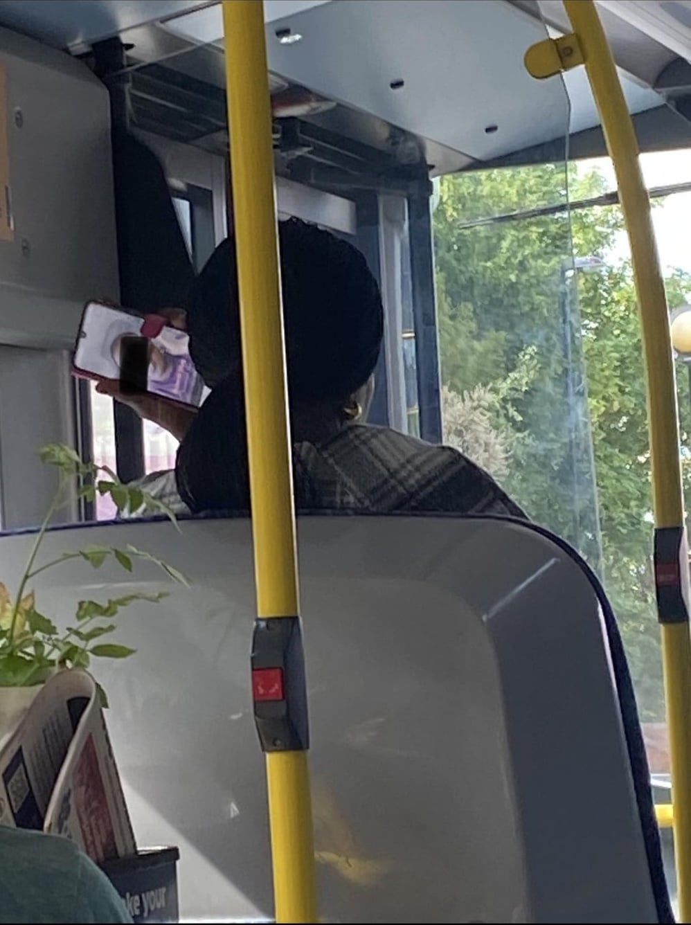 A person with dark hair sits on a bus, facing forward and holding up a phone. Outside, green trees are visible through the window. Yellow handrails and a plant are in the foreground.