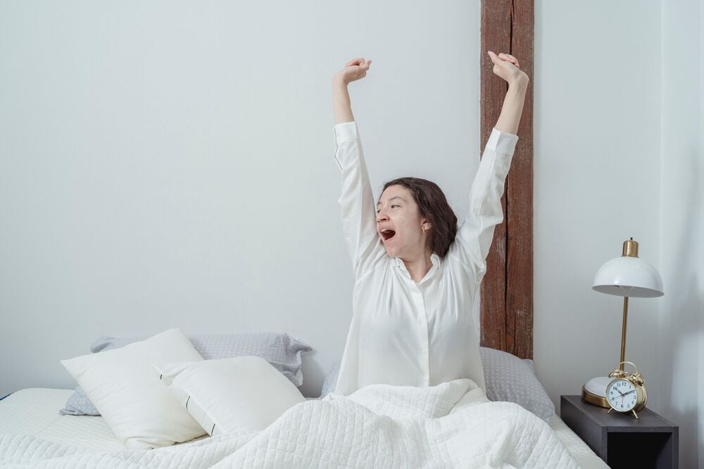 A woman in white pajamas stretches and yawns while sitting up in bed.