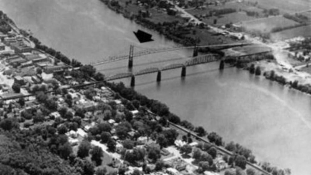 A black-and-white aerial photo shows a bridge crossing a wide river, connecting two areas with trees and buildings. An arrow points to the bridge.